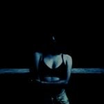 A person standing in the dark with a faint light revealing only the contours of their upper body and arms crossed, creating a moody and mysterious atmosphere.