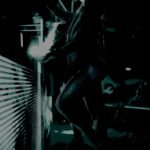 A silhouette of a person leaning against a railing at night, illuminated by a dim, atmospheric light that casts a mysterious glow.