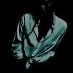 A shadowy, moody portrait of a person in a dark setting, featuring high contrast that highlights the silhouette and details of their shirt and posture.