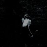 A monochromatic image captures a person with a dynamic pose against a dark brick wall, exuding an urban, artistic vibe.