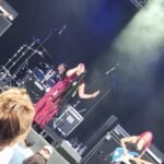 A dynamic live music performance in action, featuring a vocalist passionately singing into a microphone with a drummer and guitarist performing in the background, all under the bright stage lights in front of an engaged audience.
