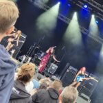 A spirited outdoor concert performance with a lead singer passionately singing on stage in a vibrant outfit, accompanied by band members, while an engaged audience watches.