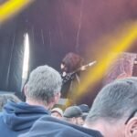 A guitarist performing on stage, viewed through a gap in the crowd at an outdoor music event.
