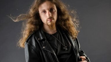 A man with long curly hair and a goatee wearing a black leather jacket against a grey backdrop, exuding a classic rocker vibe.