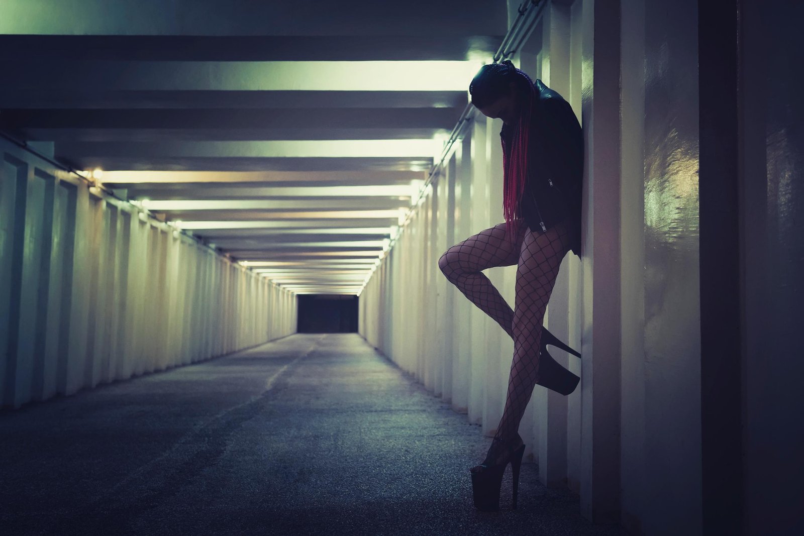 Woman in fishnet stockings and high heels leaning against a wall in a dimly lit urban tunnel, lost in thought.