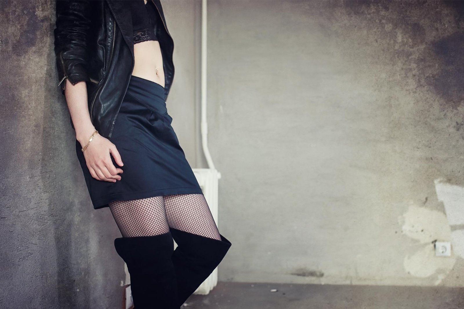 Woman in a black leather jacket and lace lingerie leaning against a rustic brick wall, gazing into the distance.