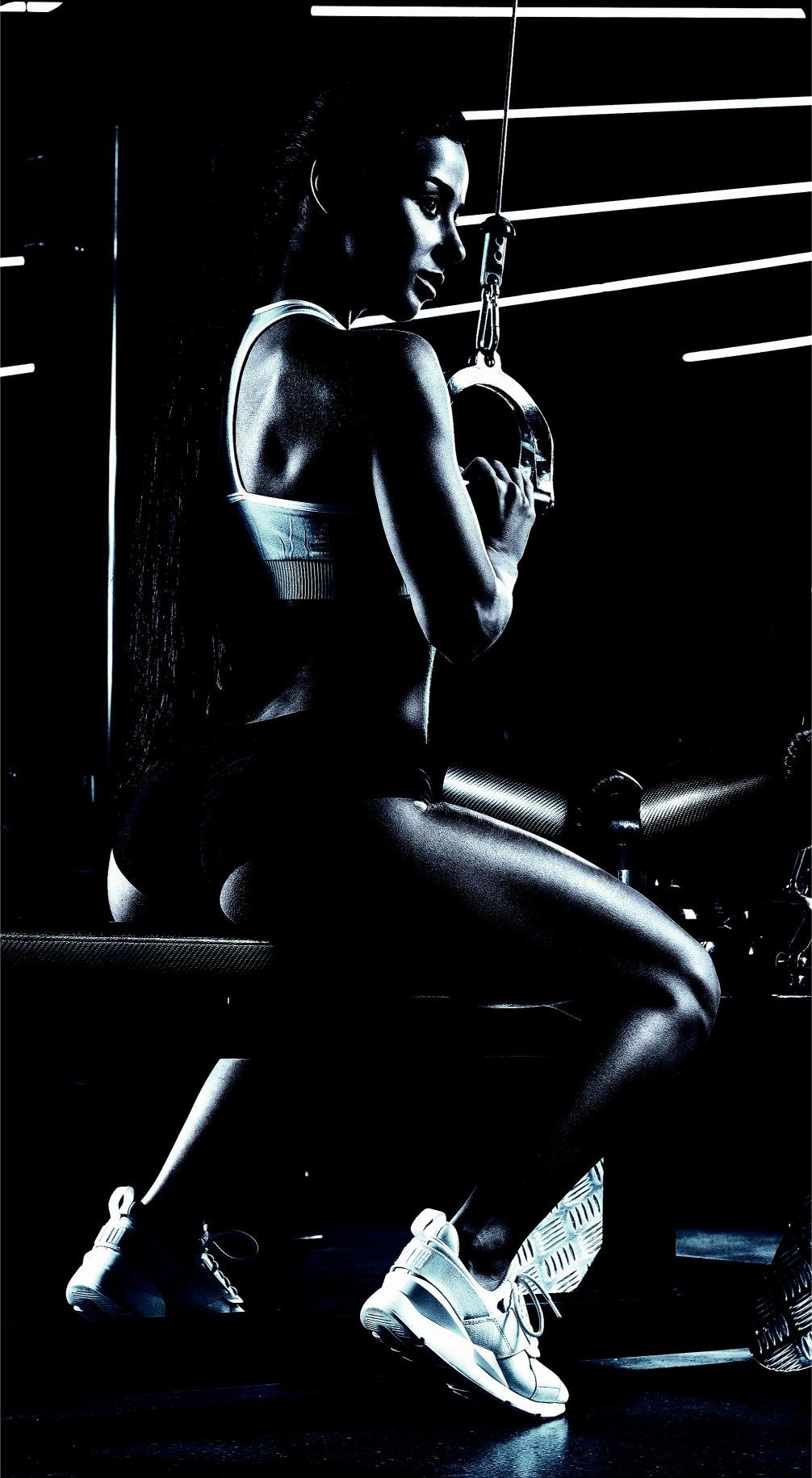 A woman seated at a gym machine in low light, gripping a handle with her back slightly turned, showcasing her toned back and arms.