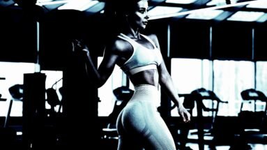 A fit woman standing in a gym, wearing a sports bra and leggings, with a strong, defined body under moody lighting.