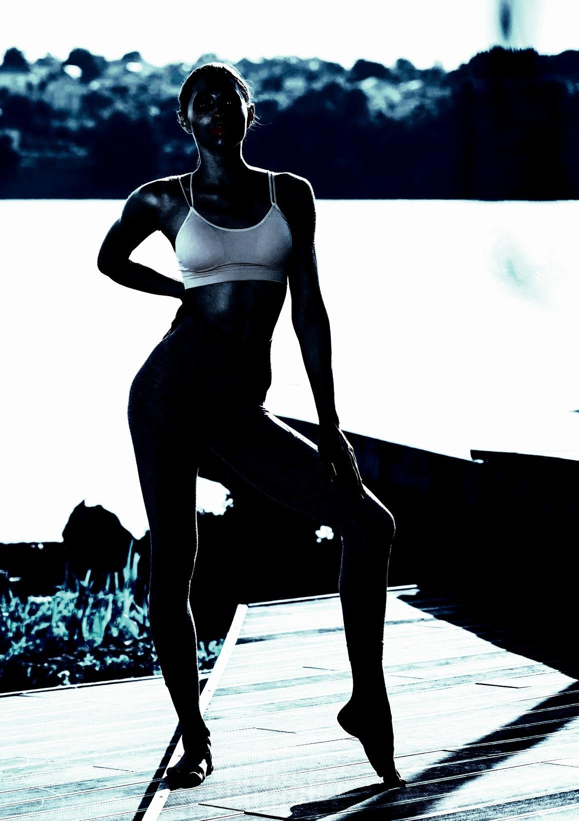 Silhouette of a woman in workout attire standing outdoors by the water, showcasing her toned body against a scenic background.