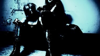 Two gothic individuals wearing rock-inspired outfits and sunglasses, sitting against a distressed wall in low blue lighting.