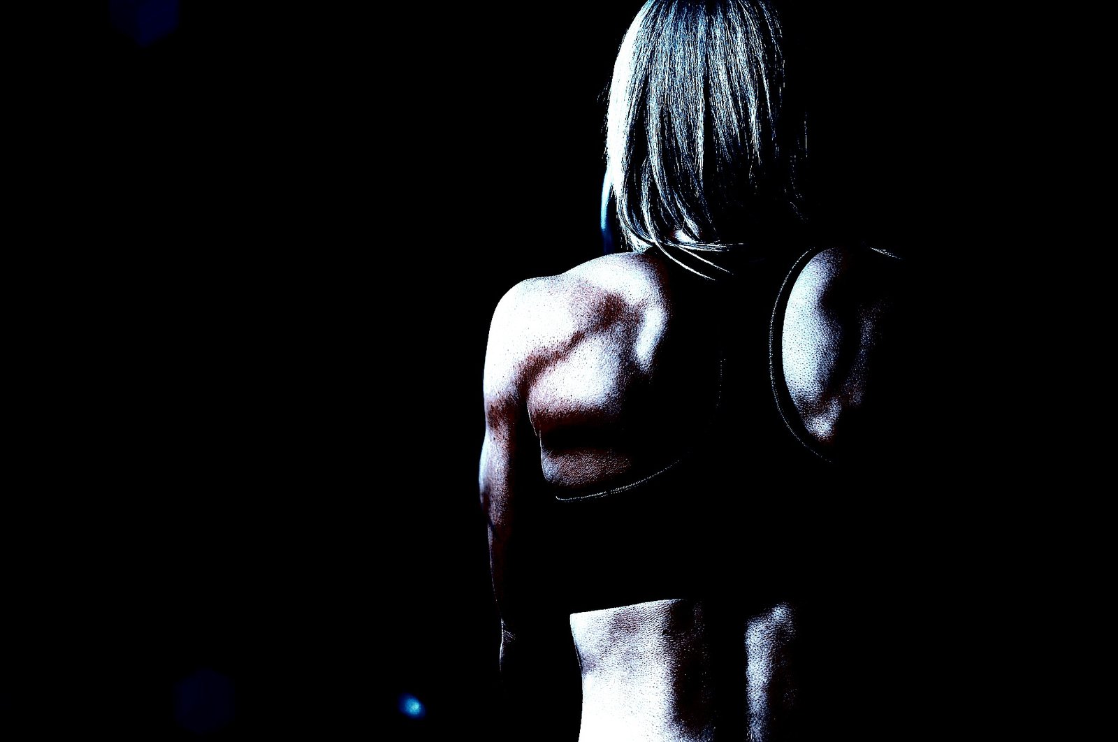 Back view of a muscular woman in a sports bra, showcasing her toned back and shoulders under low lighting.