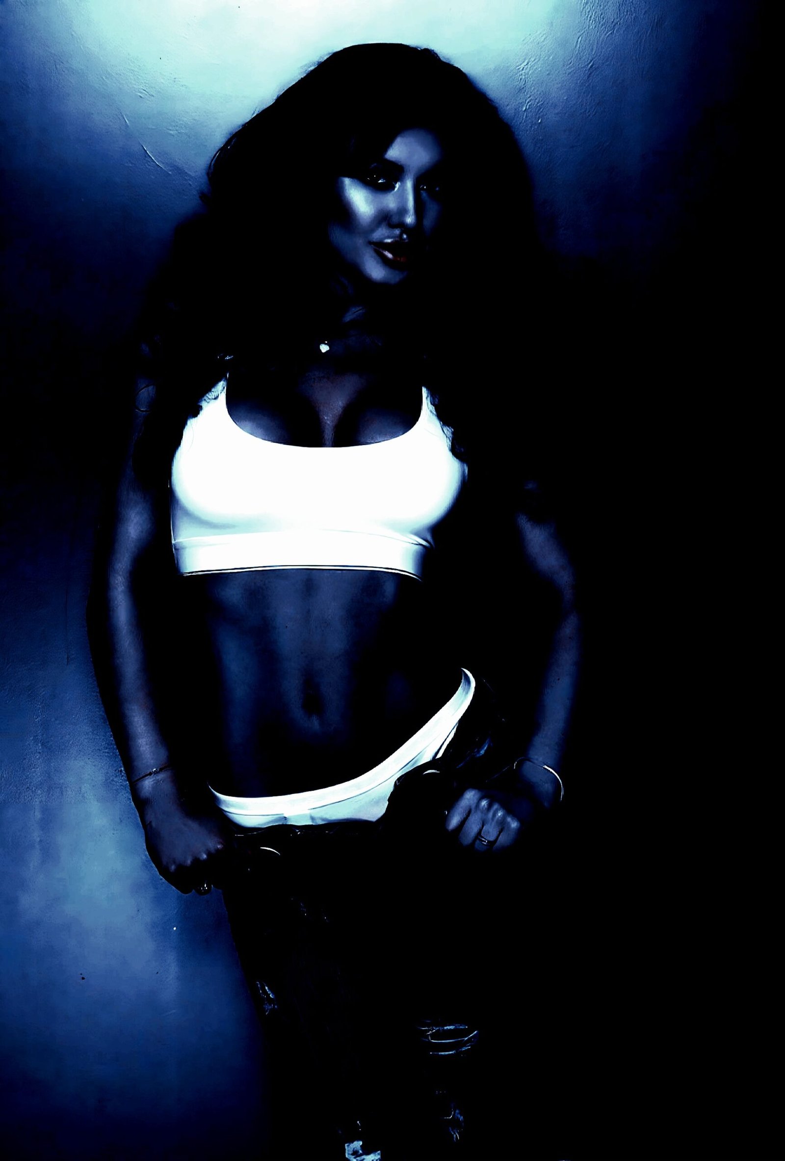 A woman in a white crop top and black pants poses against a blue wall, exuding a mix of goth allure and bold elegance.