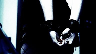 Trust and Consent in Bondage Play: A close-up black and white photo of a person's hands gently clasped together, with the wrists secured by leather cuffs, conveying vulnerability and trust.