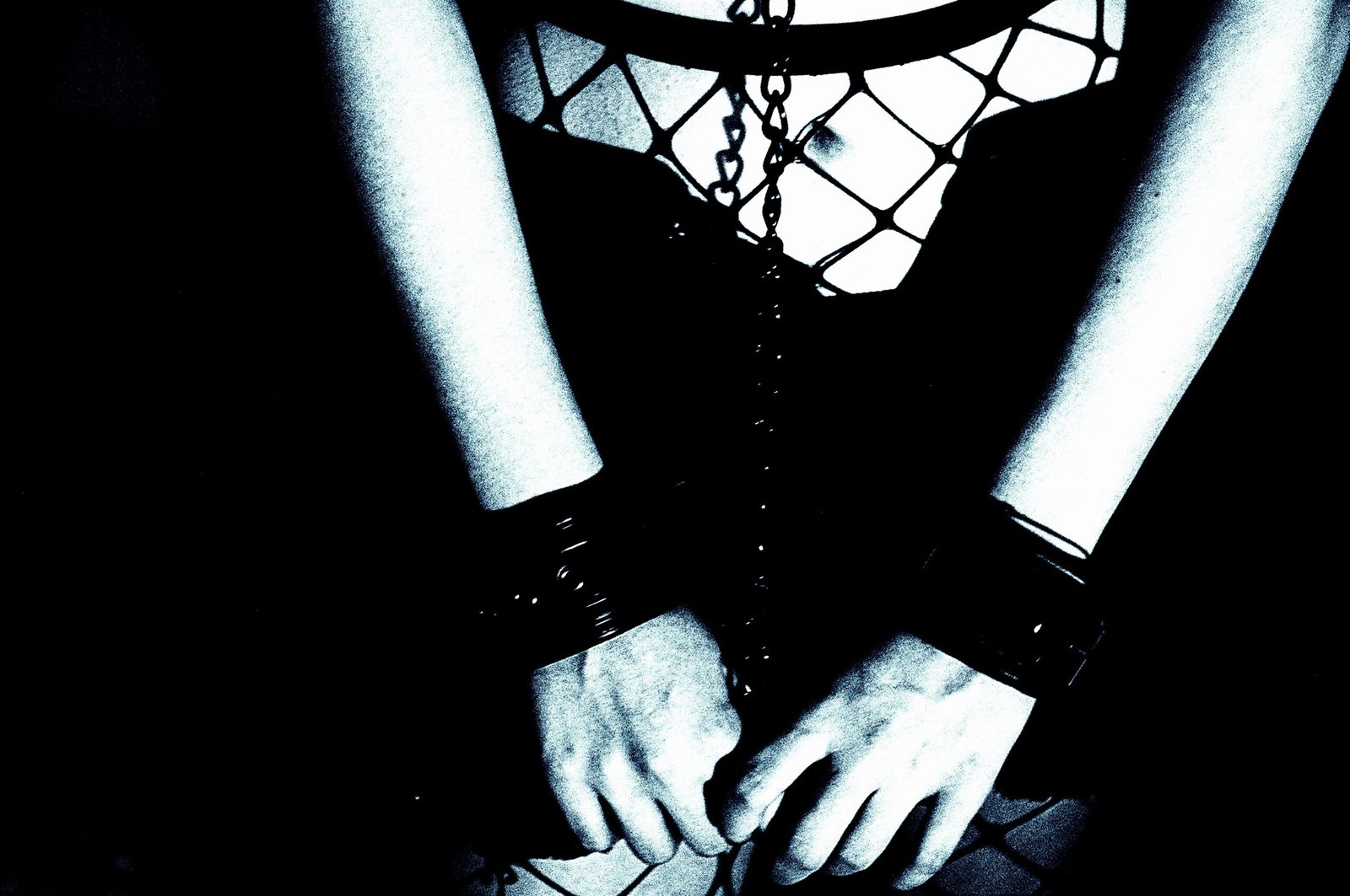 Monochrome image showcasing a person's arms bound by leather cuffs, with a hint of a spiderweb-patterned garment in the background, emphasizing the aesthetic of restraint.