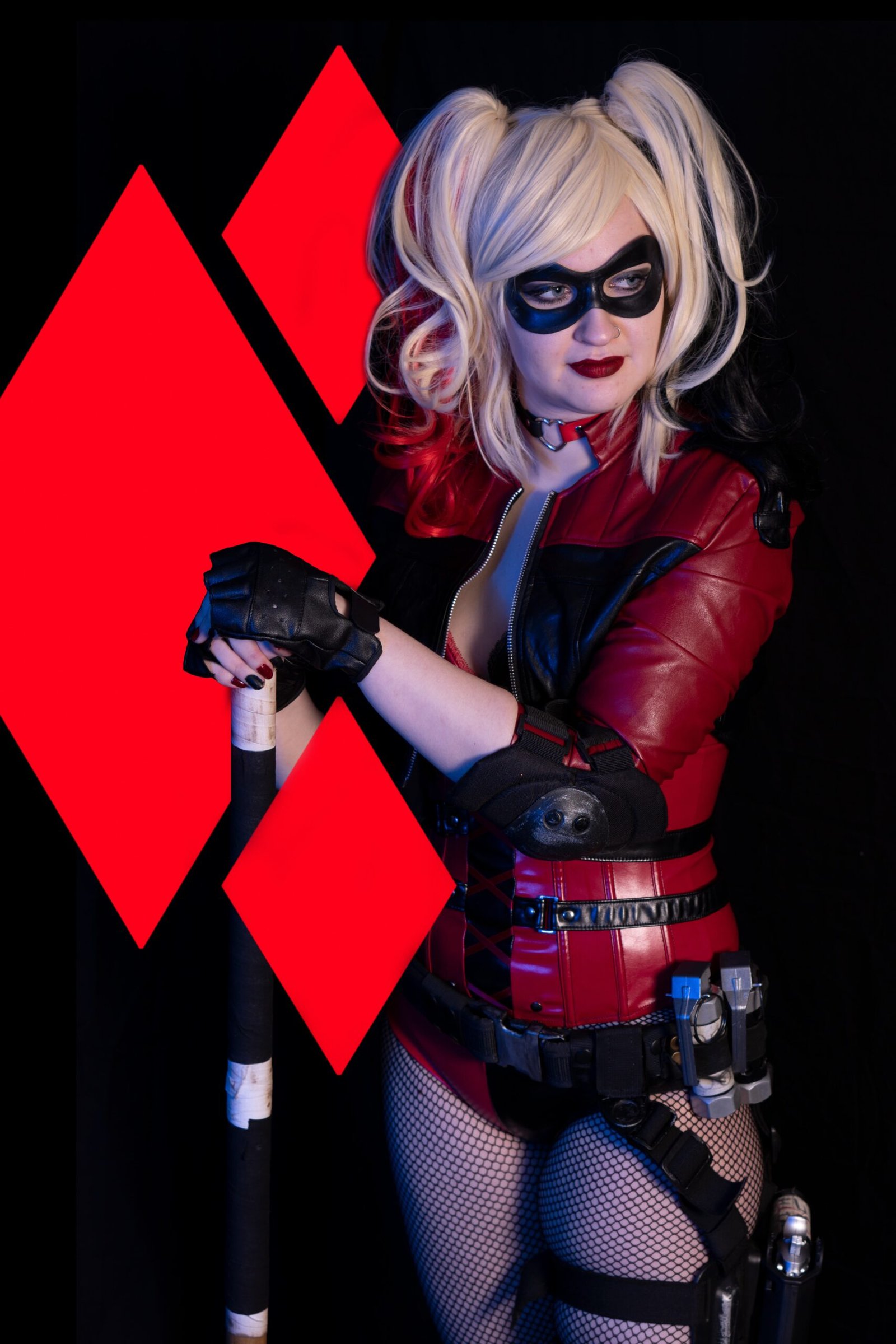 Cosplayer as a comic book anti-heroine in a red and black costume with a bat weapon, against a red diamond background.