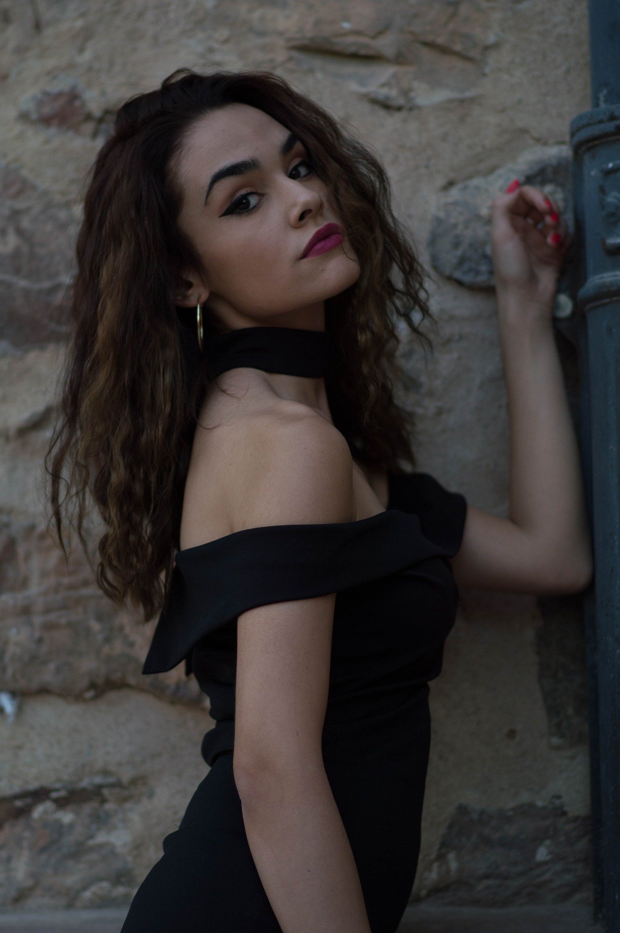 A young woman in a black off-shoulder top with a choker gives a sultry look to the camera, her wavy brown hair and makeup accentuating her soft features as she leans against a weathered stone wall.