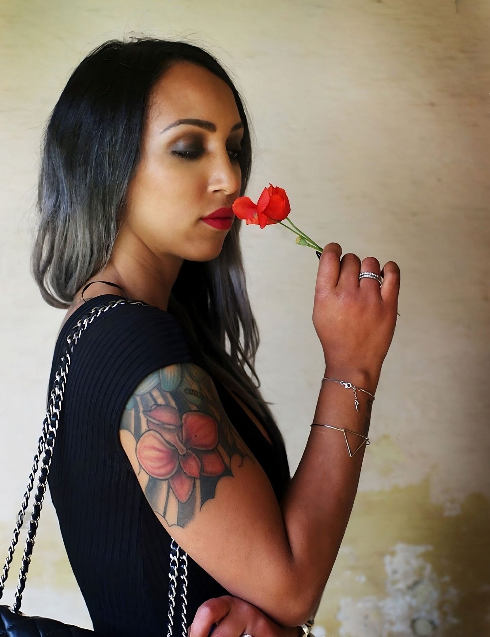 Woman with ombre hair and a sleeve tattoo smelling a red rose, wearing a black dress and silver bracelets.