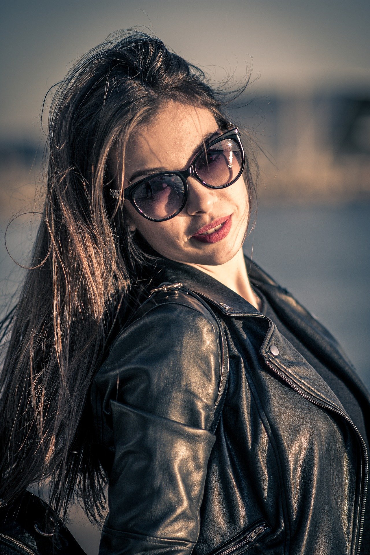 Close-up of a woman in sunglasses and leather jacket, with a pensive expression and windblown hair.