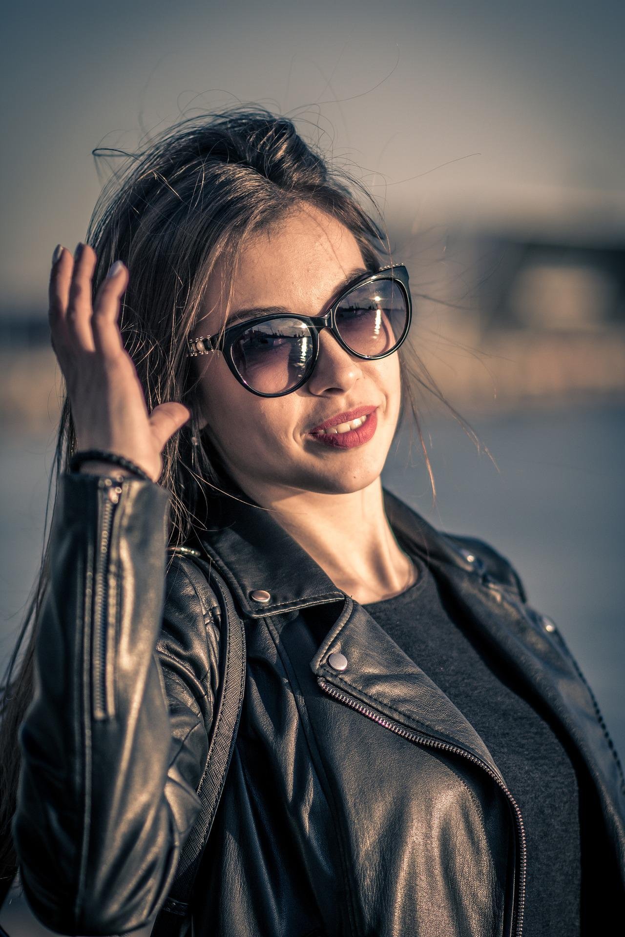 Stylish woman touching her sunglasses, wearing a black leather jacket with a softly lit background.