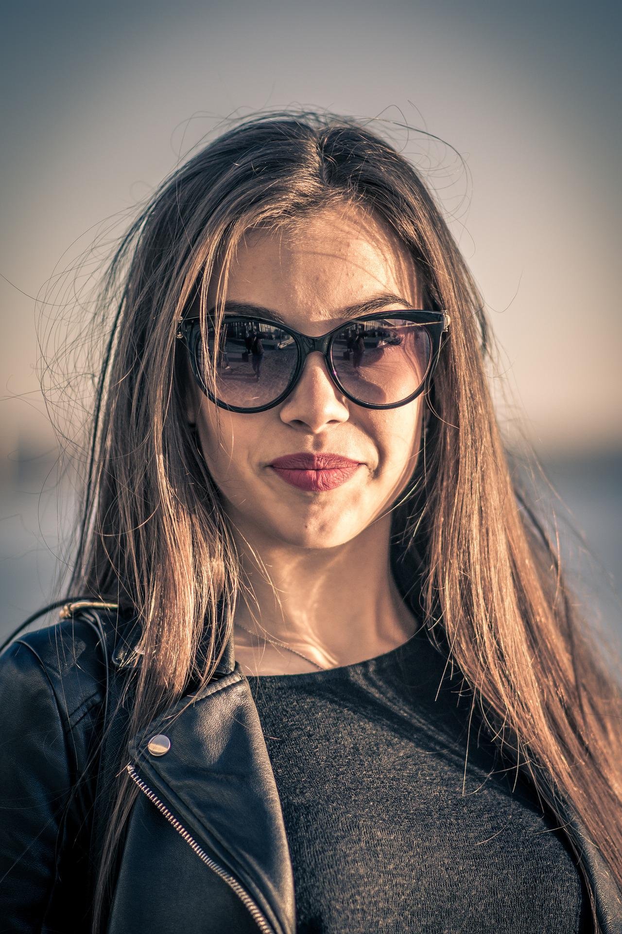 Woman in a casual pose wearing a black shirt and leather jacket, large sunglasses, and a confident half-smile.