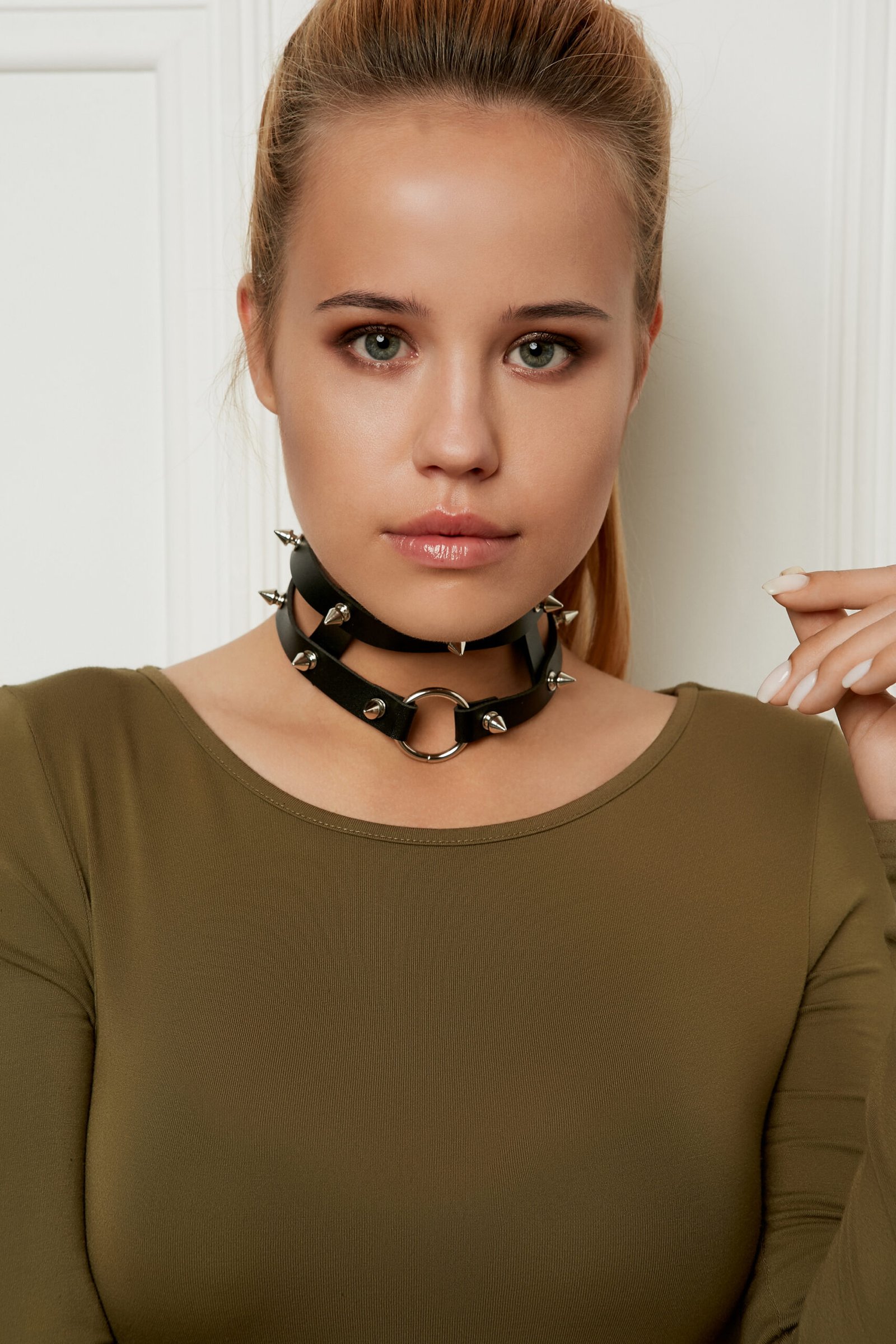 A woman wearing a spiked choker and an olive green top, looking pensively to the side.