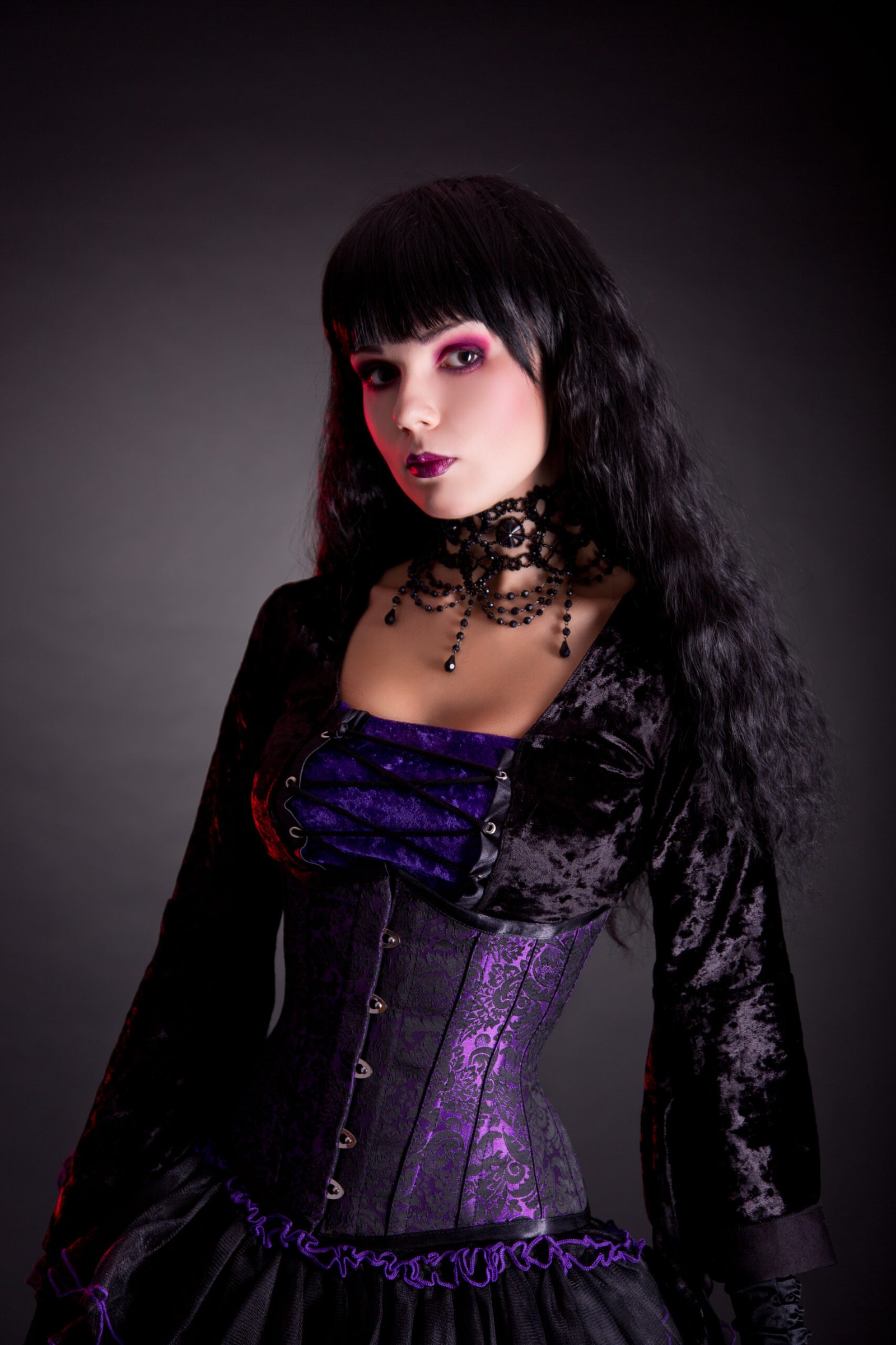 A woman in gothic attire standing straight with a slight tilt of the head, her layered choker necklace and intricate corset detail in focus against the dark backdrop.