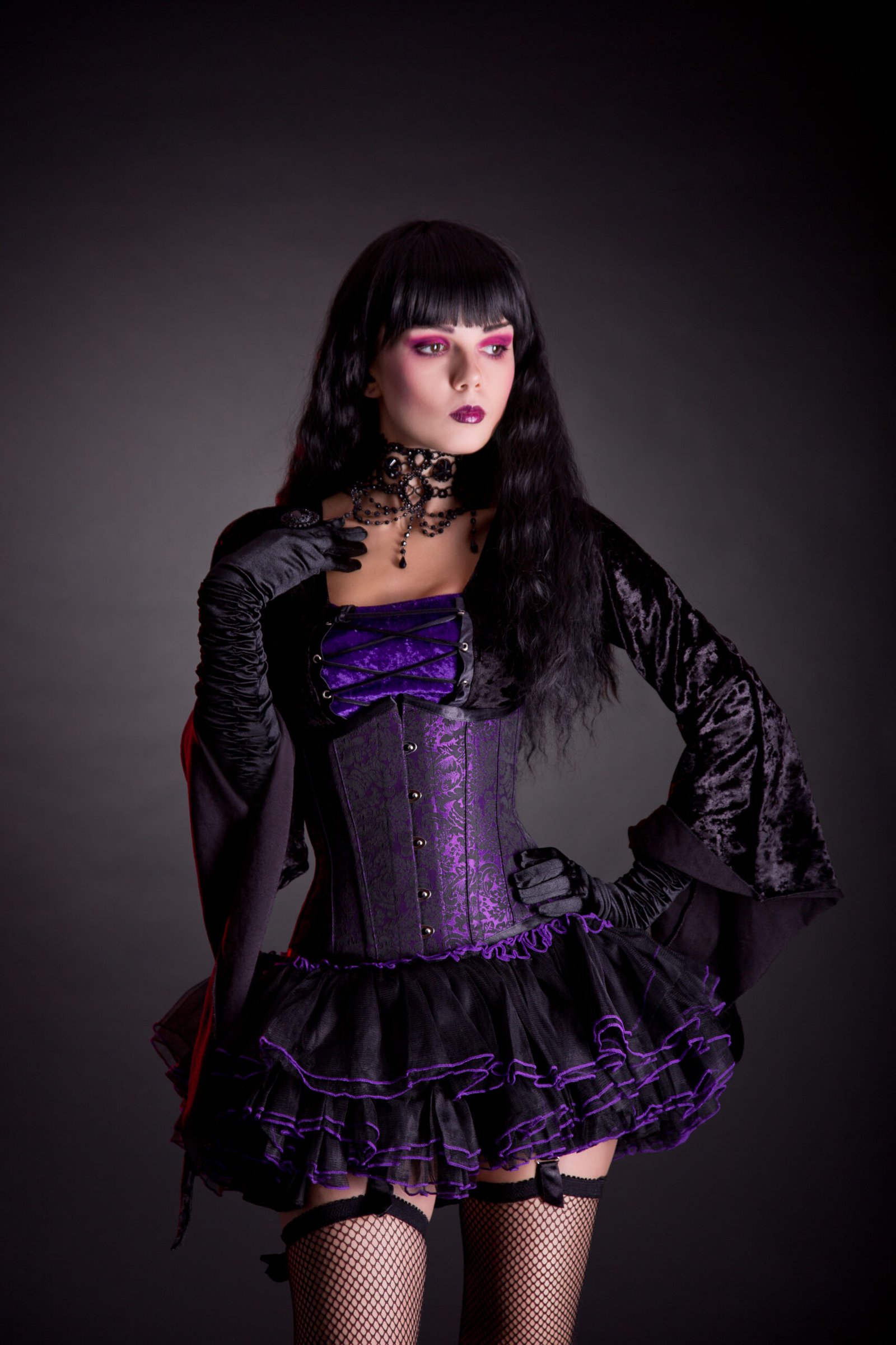 Woman in full gothic regalia holding an ornate black umbrella, her striking makeup, and choker necklace accentuating the dark, romantic aesthetic of her outfit.