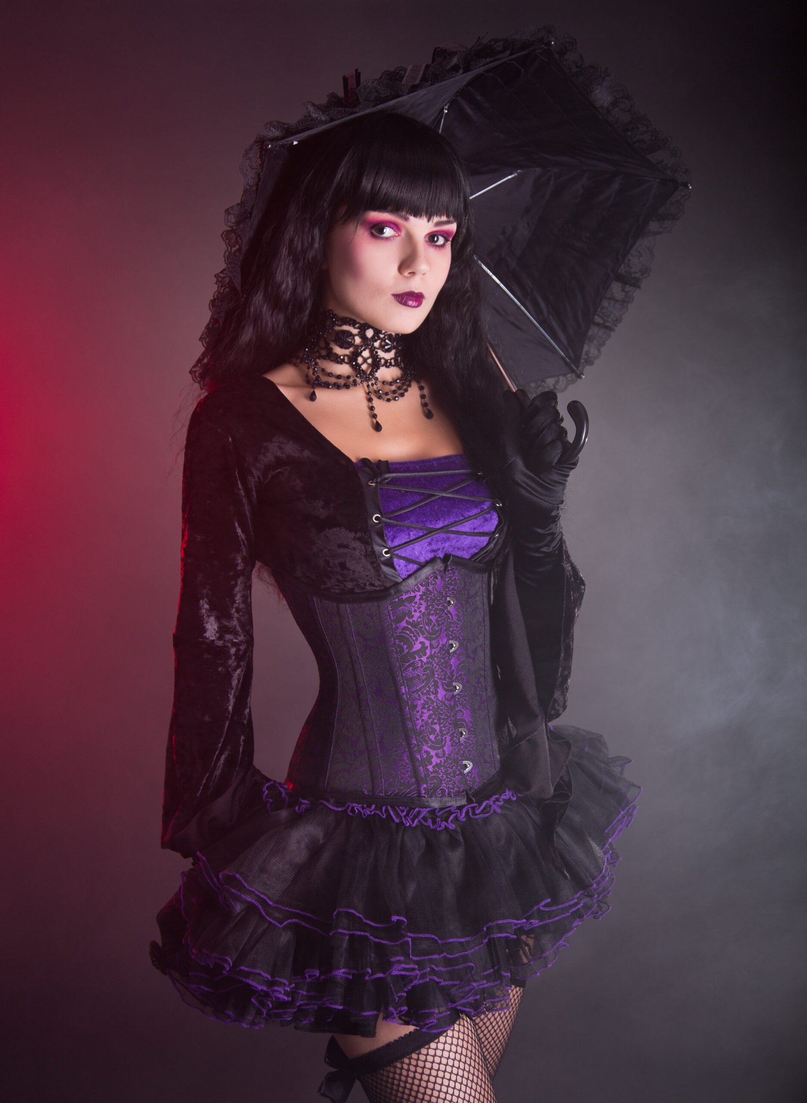 The goth woman is poised with an umbrella over her shoulder, her black velvet jacket and layered skirt highlighted against the misty red-tinged background.
