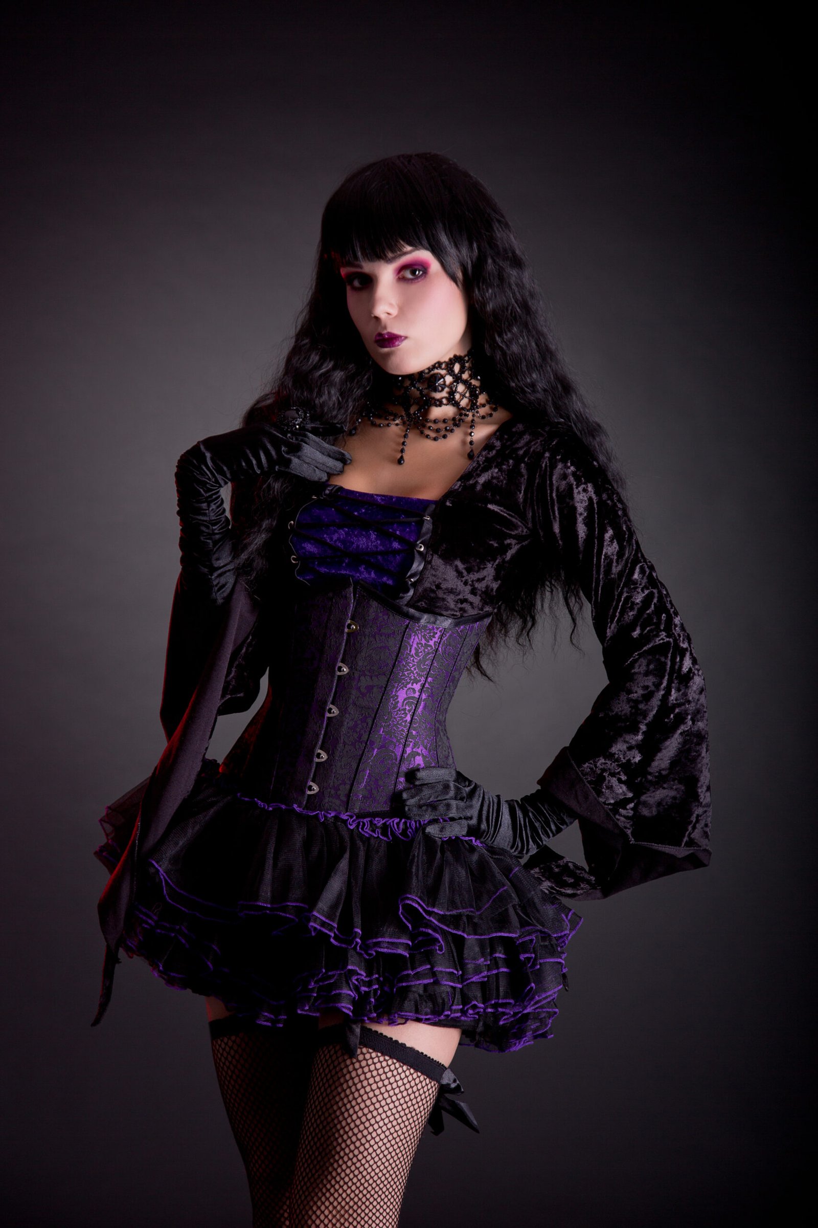 A woman with long black hair wearing a goth-inspired outfit with a purple and black corset, black velvet jacket, ruffled skirt, and fishnet stockings, poised in a dramatic stance against a dark background.