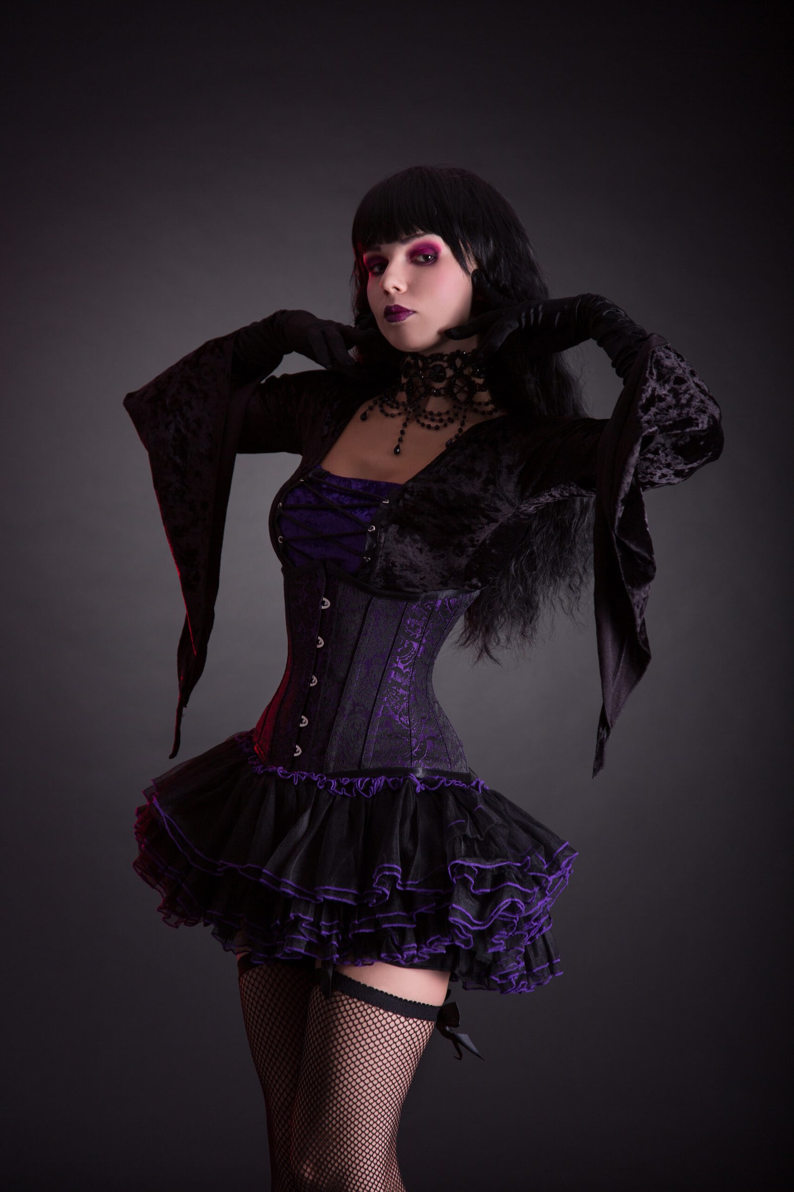 Goth woman striking a confident pose with one hand on her waist, her black velvet jacket's sleeve draping elegantly, complementing her corset and skirt ensemble.