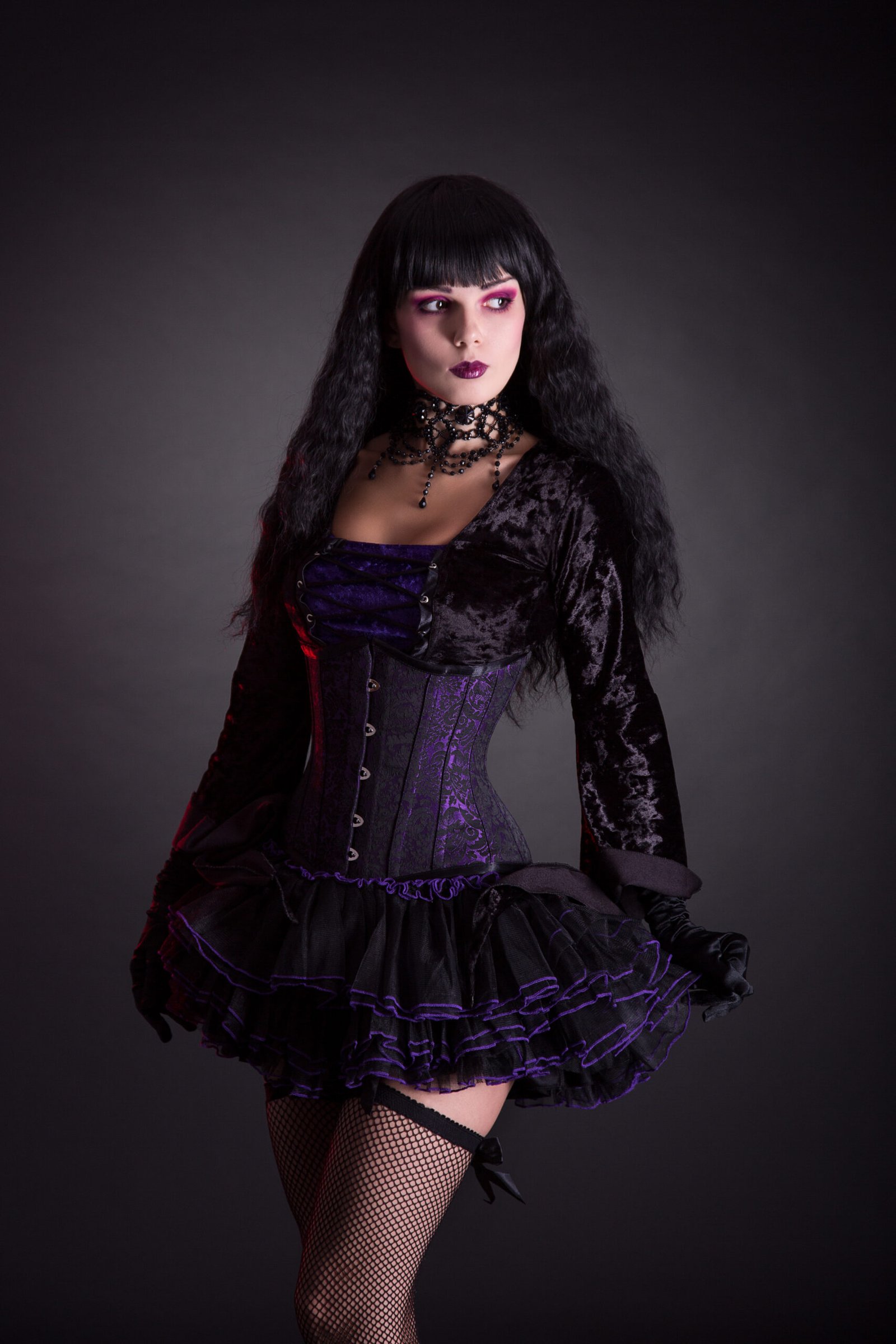 Woman in a gothic costume holding a black top hat in one hand, standing confidently with her other hand on her hip, showcasing her vibrant makeup and layered choker.