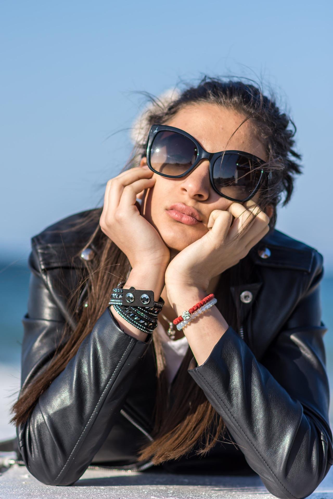 Woman lounging with head resting on hands, wearing oversized sunglasses and black leather jacket, adorned with beaded bracelets.
