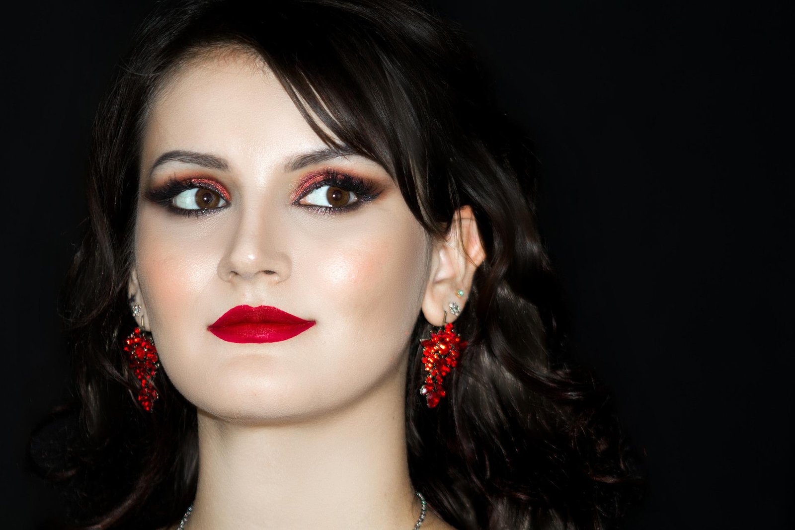 A woman with vivid makeup, including smoky eyes and bold red lipstick, exudes a glamorous vibe with sparkling red earrings as she poses against a stark black background.