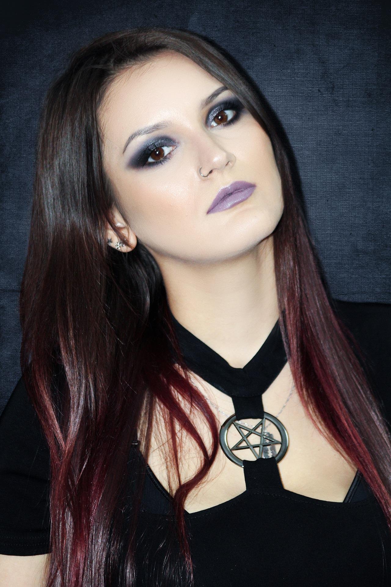 Portrait of a woman wearing dark makeup, with her eyes outlined in black, sporting a black top with a pentagram necklace; her hair is a mix of purple and black, indicative of a goth aesthetic.