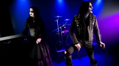 Eerie stage scene with a gothic duo, woman in black lace and man in spiky attire, playing synths in a foggy, dimly lit setting. Ashbury Heights - SmALLer.