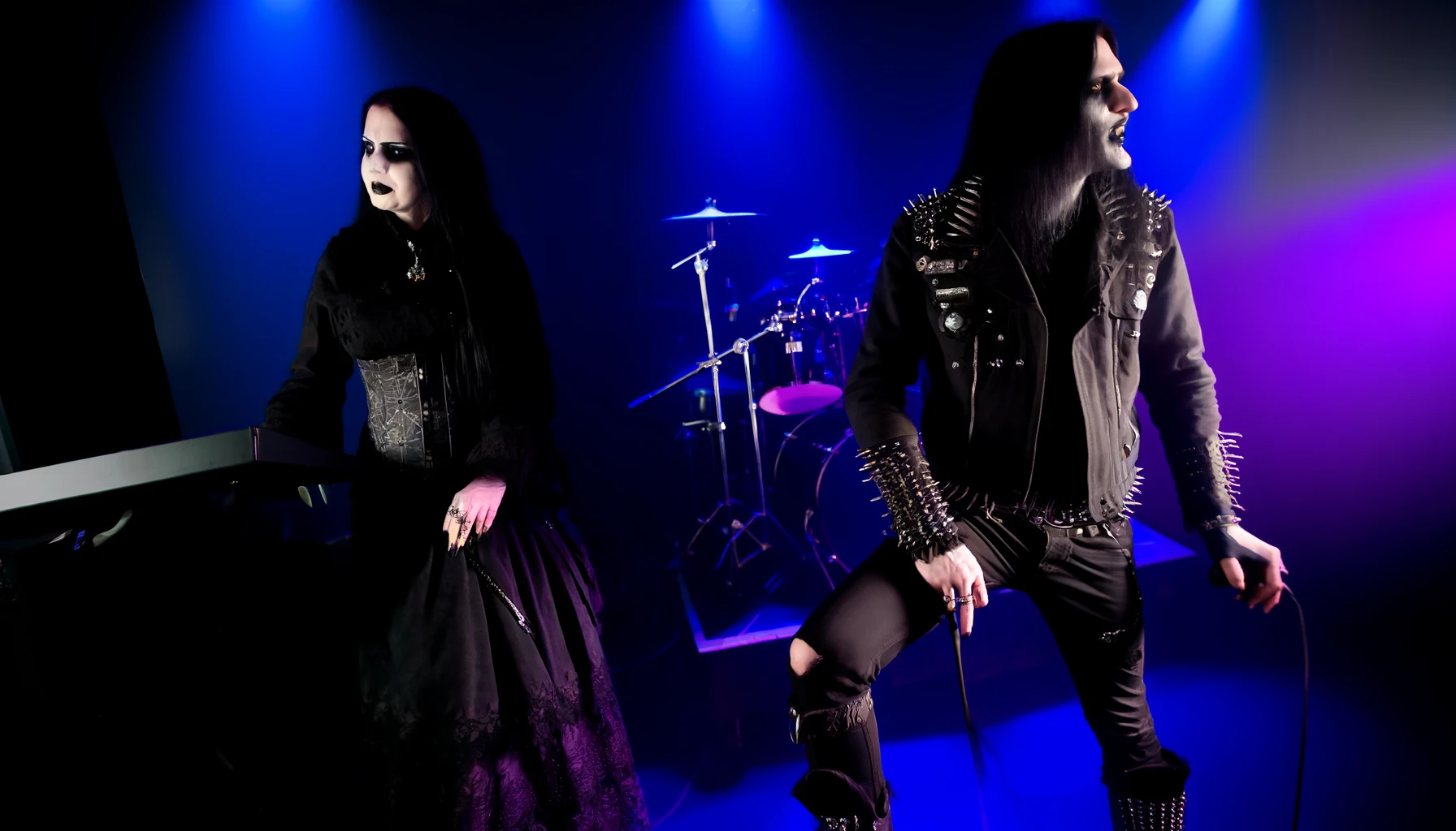 Gothic duo on stage with synthesizers, featuring a woman in elaborate black dress and a man in studded leather, both with intense dark makeup, under moody blue and purple lighting. Ashbury Heights - SmALLer.