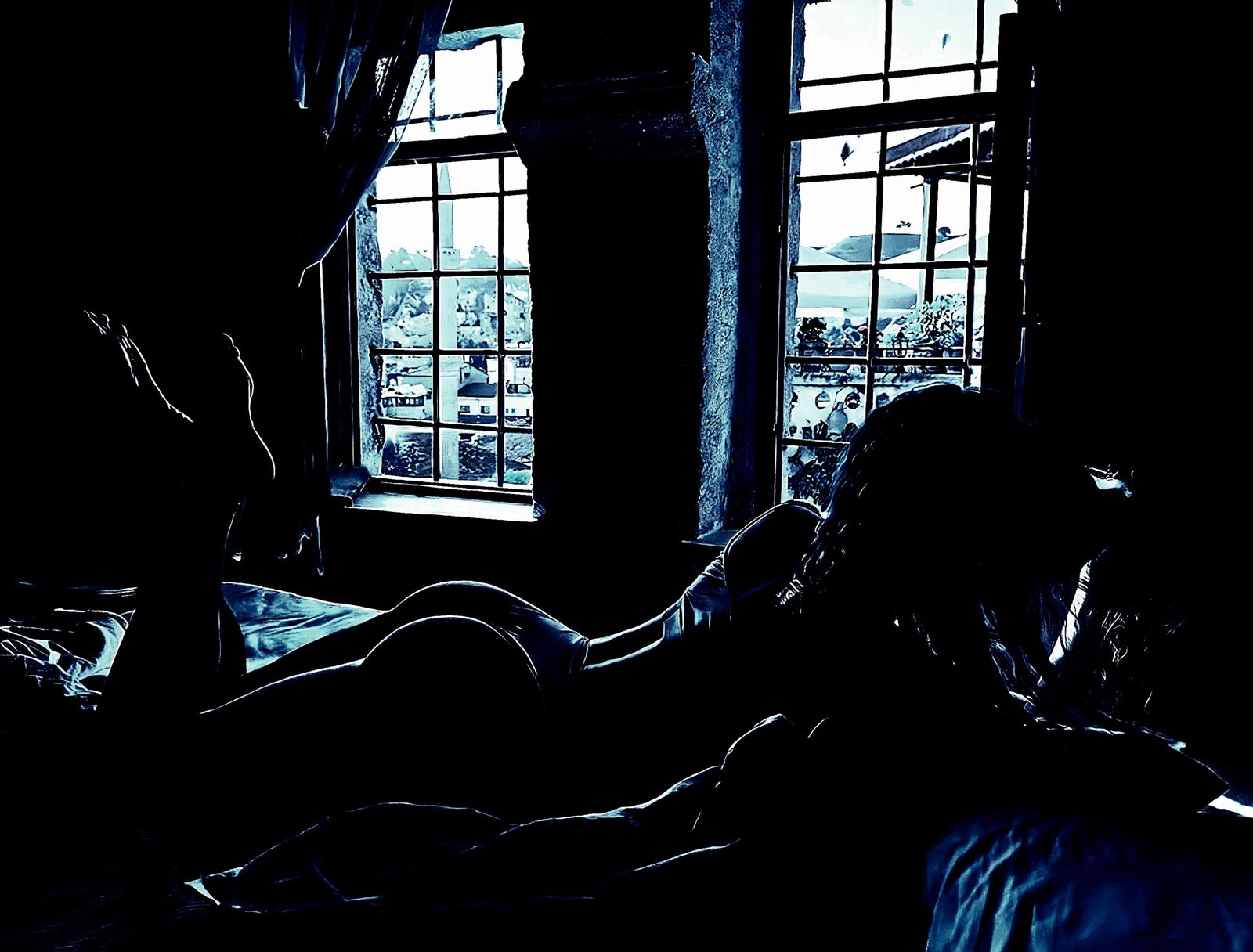 Silhouette of a person lying on a bed in front of large windows, with a view of an urban landscape outside.