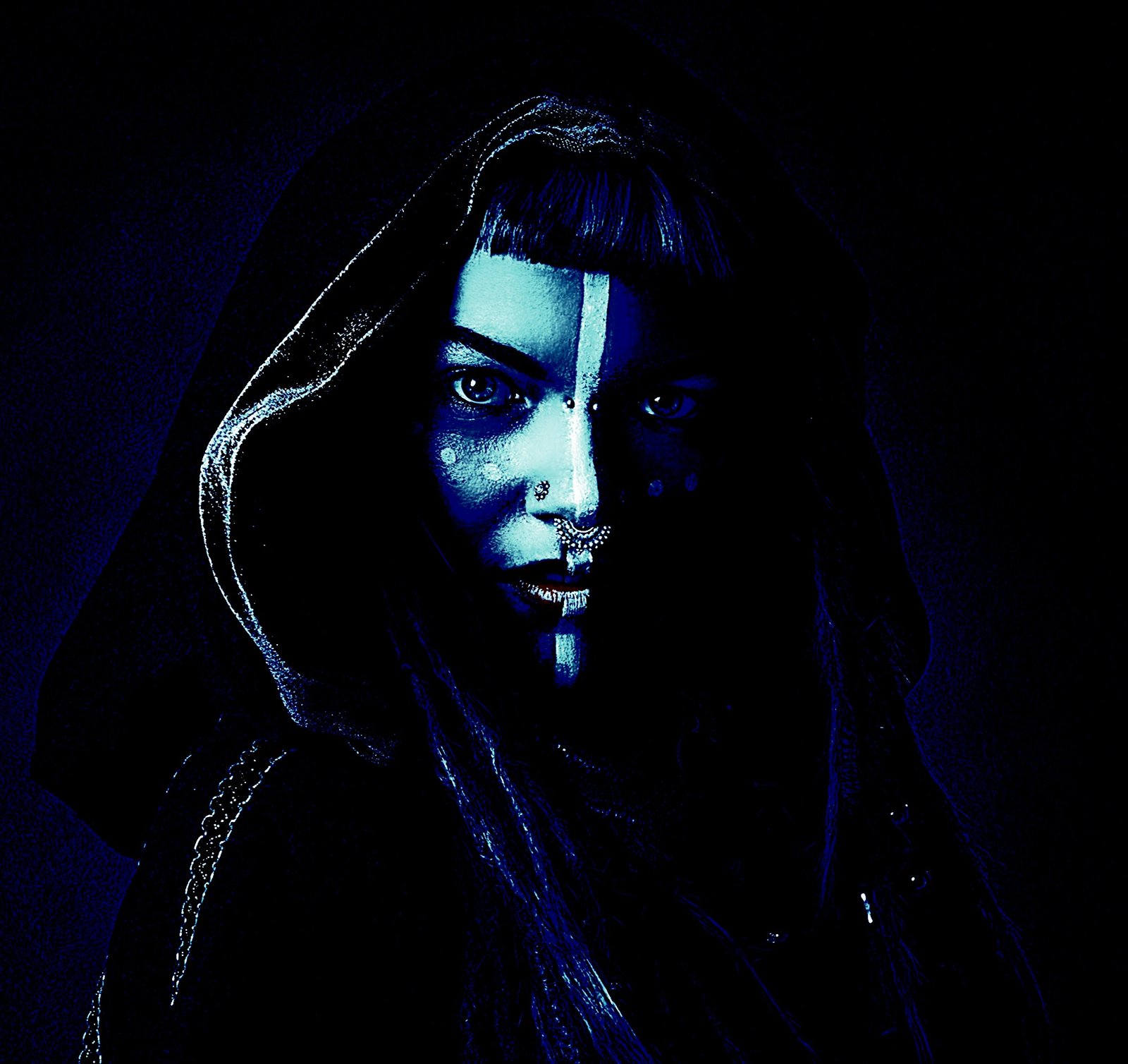 Intense gothic makeup with blue hues on a woman wearing a hood, featuring facial piercings and mystical face paint.