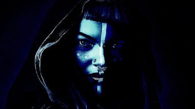 Wide shot of a woman in dark gothic attire and face paint, standing enigmatically in a dimly lit room.