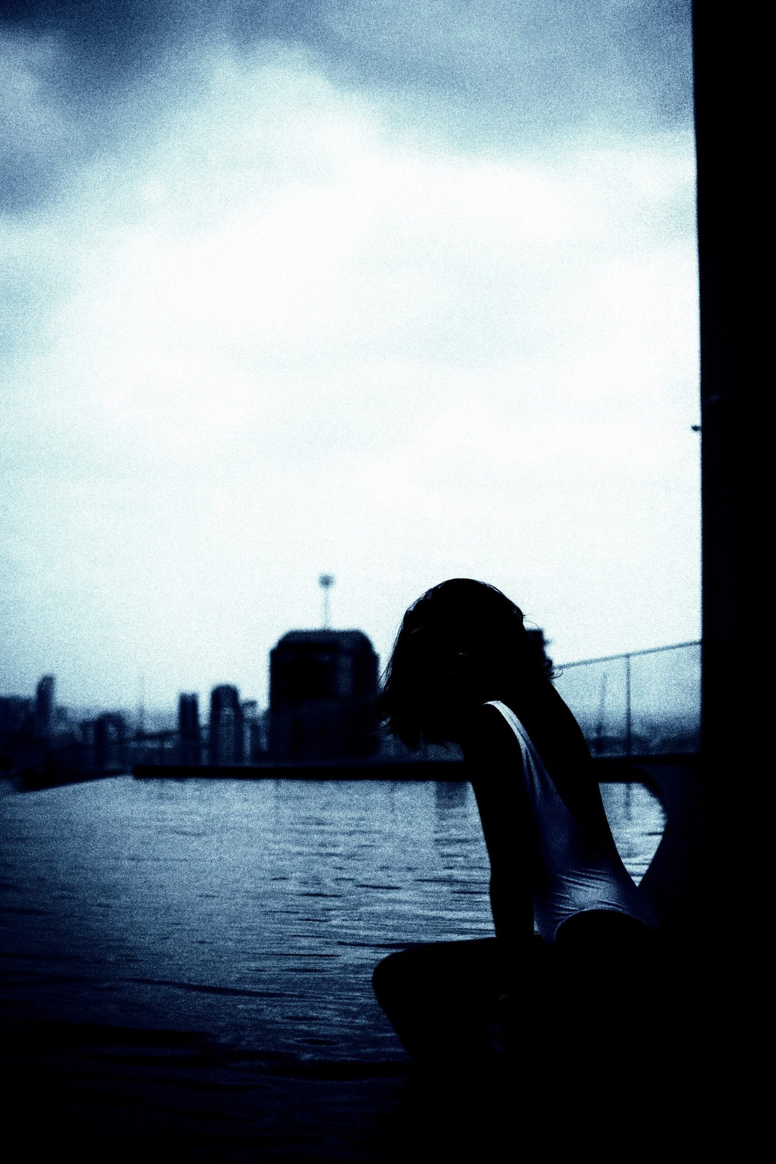 Silhouette of a person sitting by the edge of a rooftop pool at dusk, with a cityscape in the background.