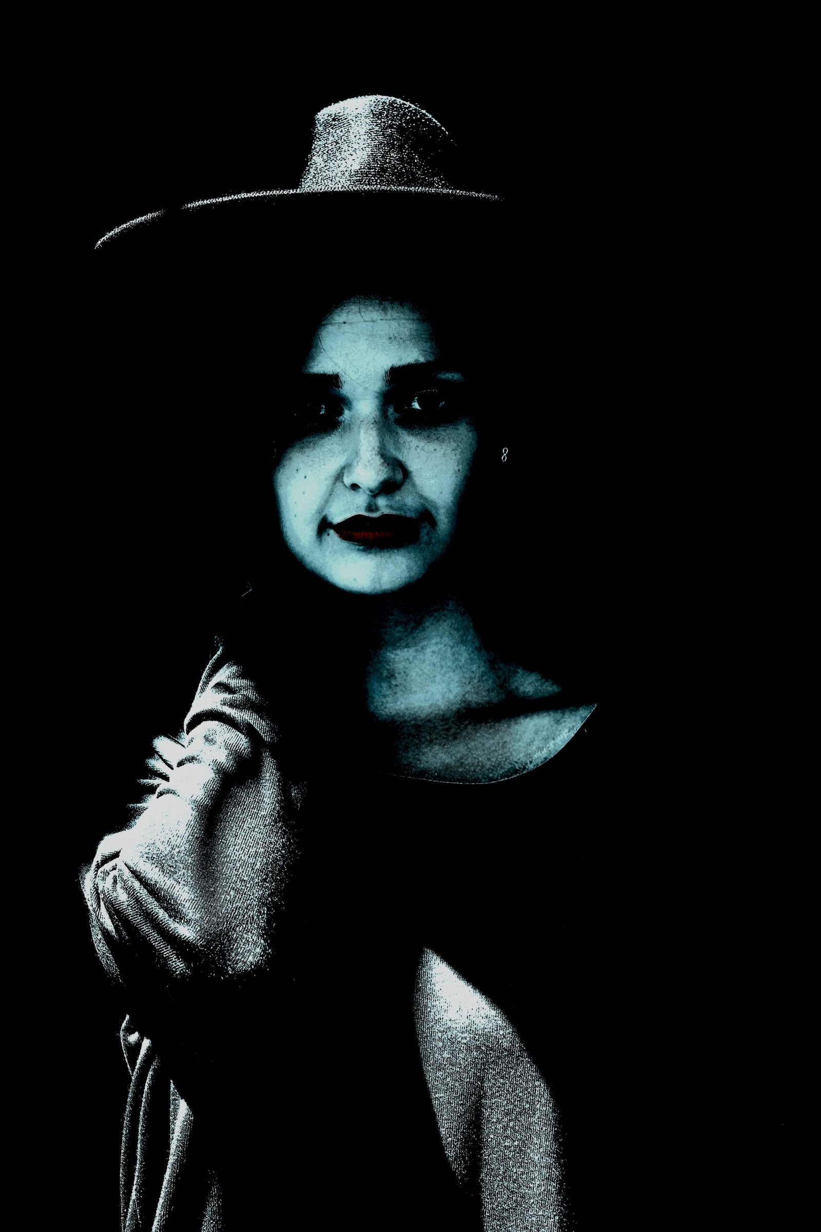 Woman in a dark outfit and wide-brimmed hat, with a somber expression under stark blue lighting.