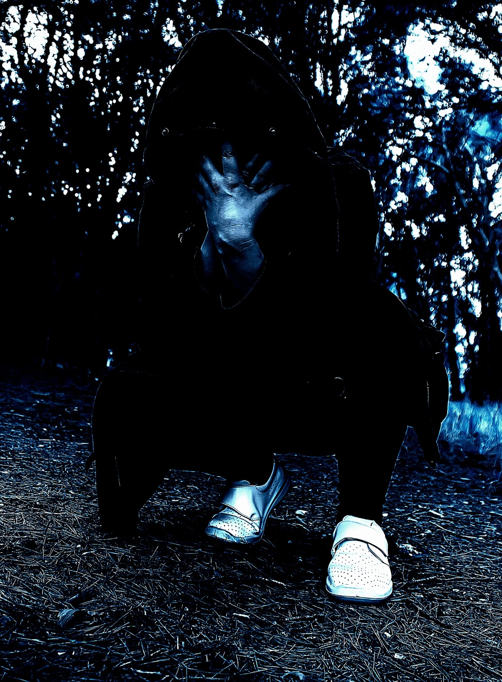 Shadowy figure in a gothic outfit hiding face with hands, set against a background of blurred forest foliage.
