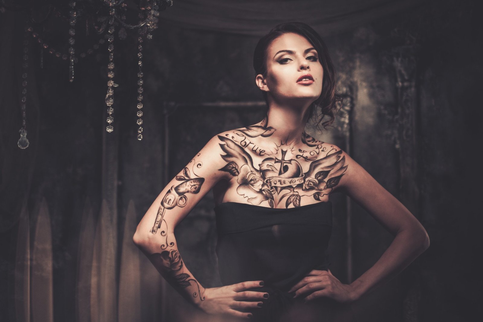 Elegant tattooed woman in a dark setting with intricate chest and arm tattoos, a deep look into Tattoos in the Metal Subculture.