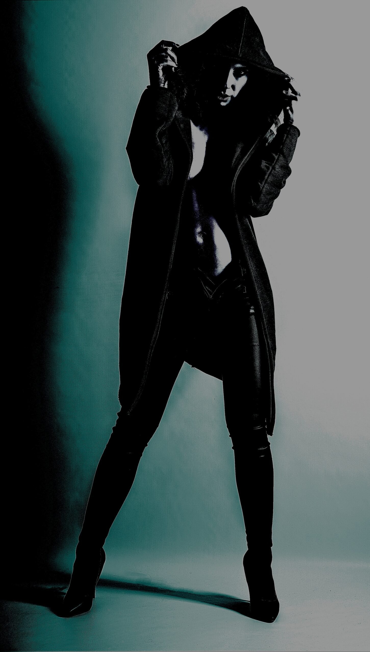 Gothic fashion model in a black hooded coat and boots, posing dramatically under blue lighting.