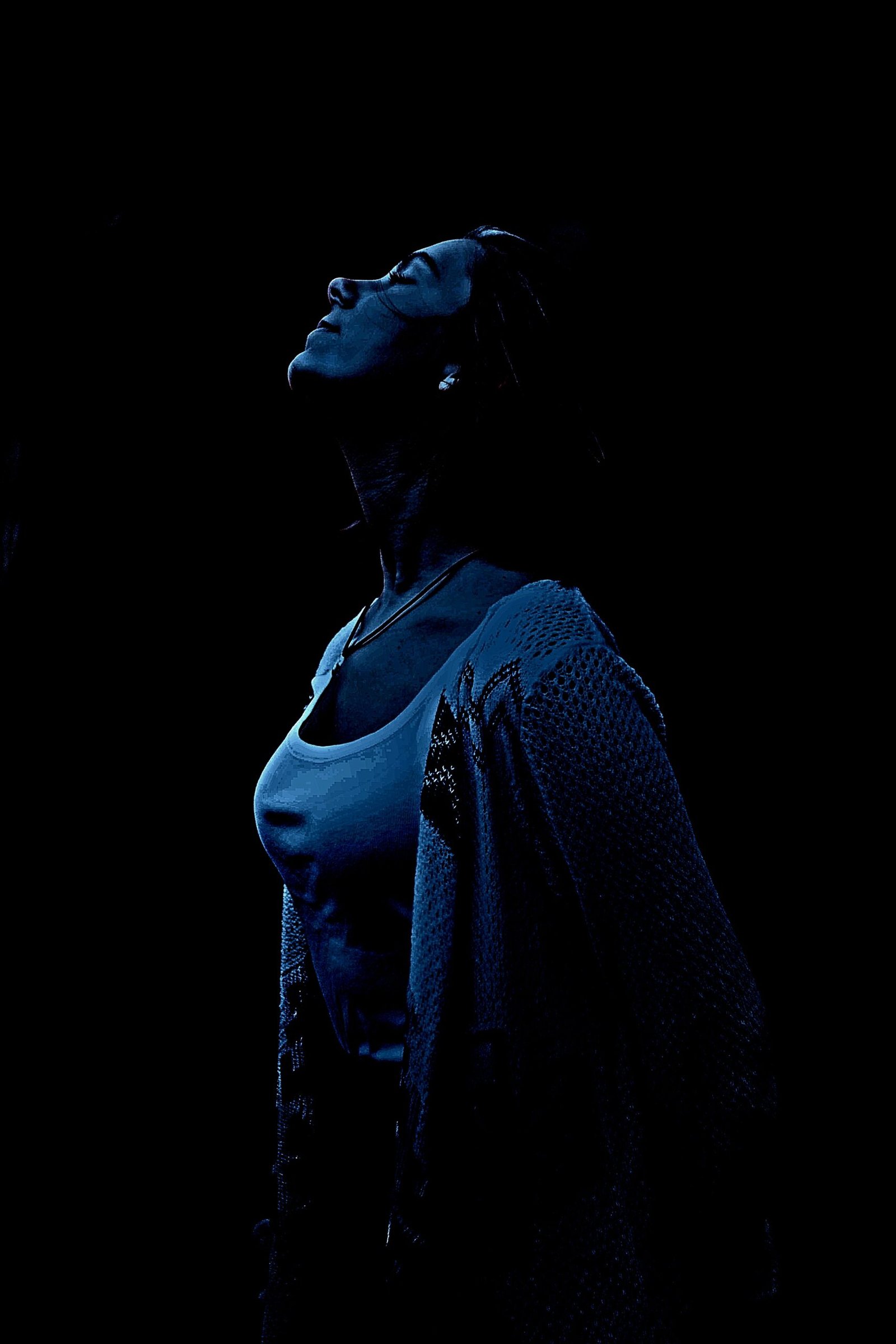 Serene pose of a woman dressed in a blue tank top and black knit cardigan, head tilted back in dim blue lighting.