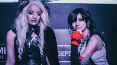 Two cosplayers posing at a gaming event, one in a dark fantasy outfit with long white hair and the other in a sporty costume with red gloves.