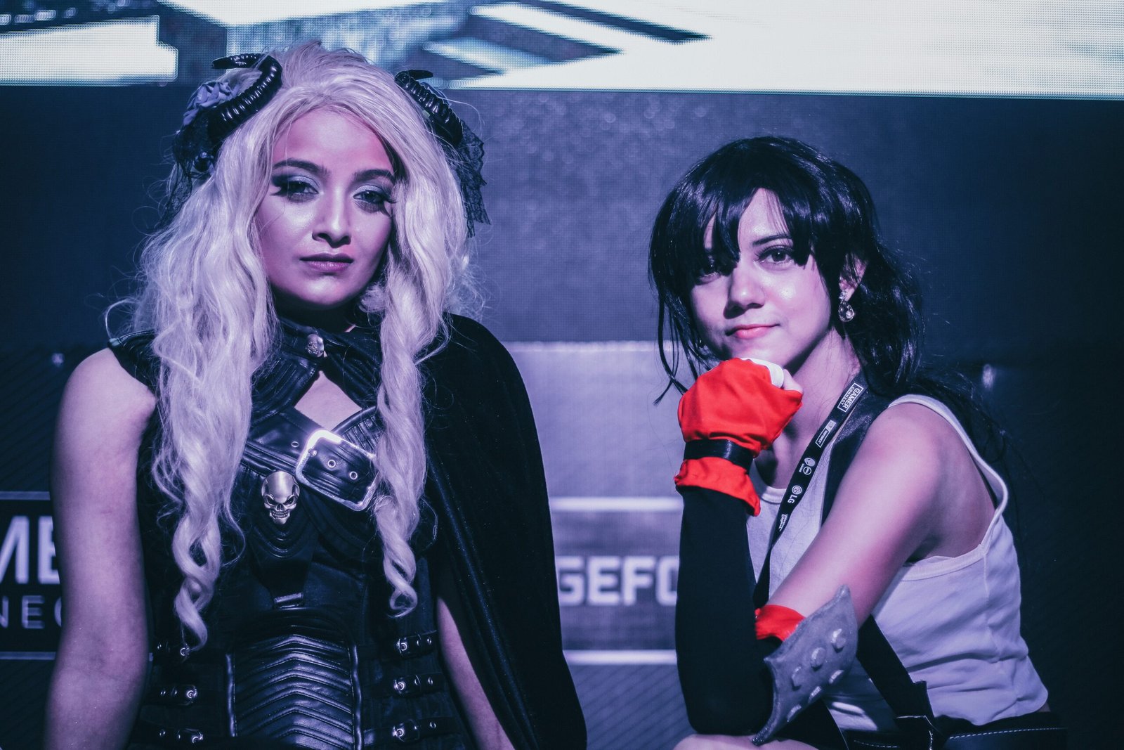 Two cosplayers posing at a gaming event, one in a dark fantasy outfit with long white hair and the other in a sporty costume with red gloves.