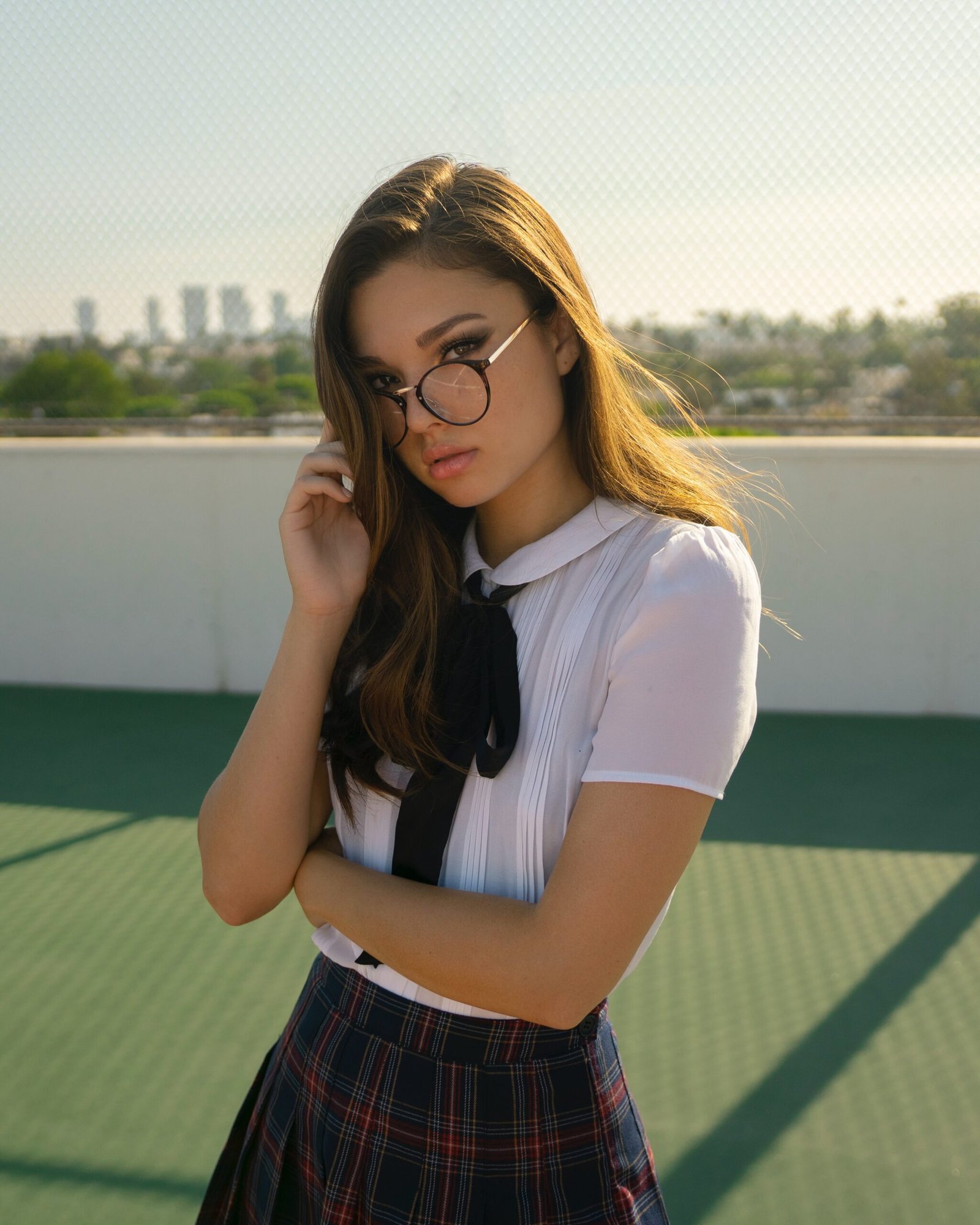 Young woman in a schoolgirl cosplay outfit, wearing glasses and standing on a rooftop with a cityscape in the background.