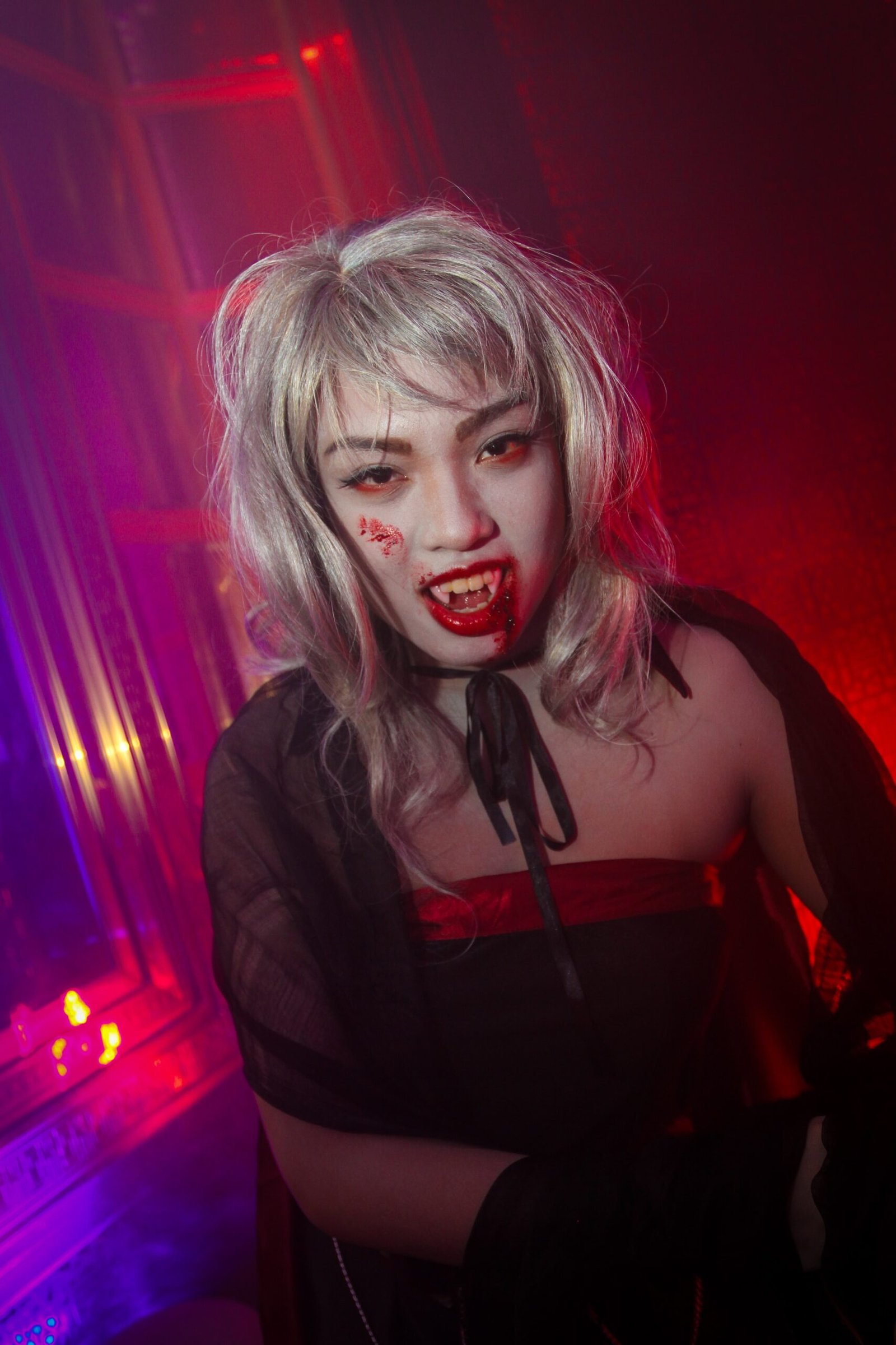 Cosplayer dressed as a vampire with silver hair and fangs, standing in a dimly lit room with red lighting.