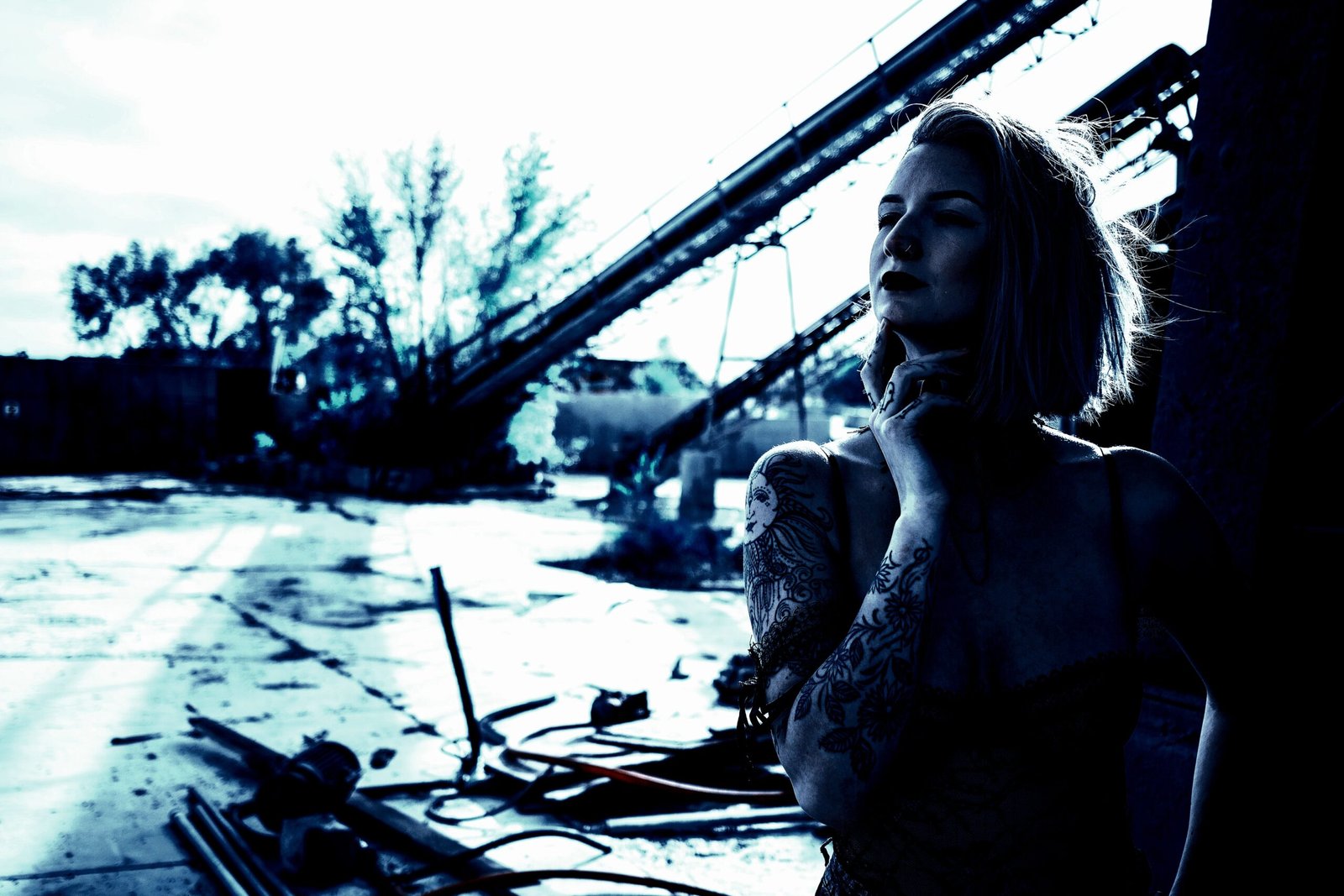 Woman with short hair and tattoos in a pensive pose against an industrial background